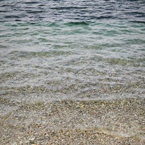 Blick vom Feinsteinufer ins klare Wasser eines Badesees. Türkisblau wird in der Ferne und Tiefe zu Dunkelblau.