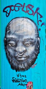 Graffiti: Kopf in Vorderansicht mit Bart und Nasenring vor hellblauem Hintergrund