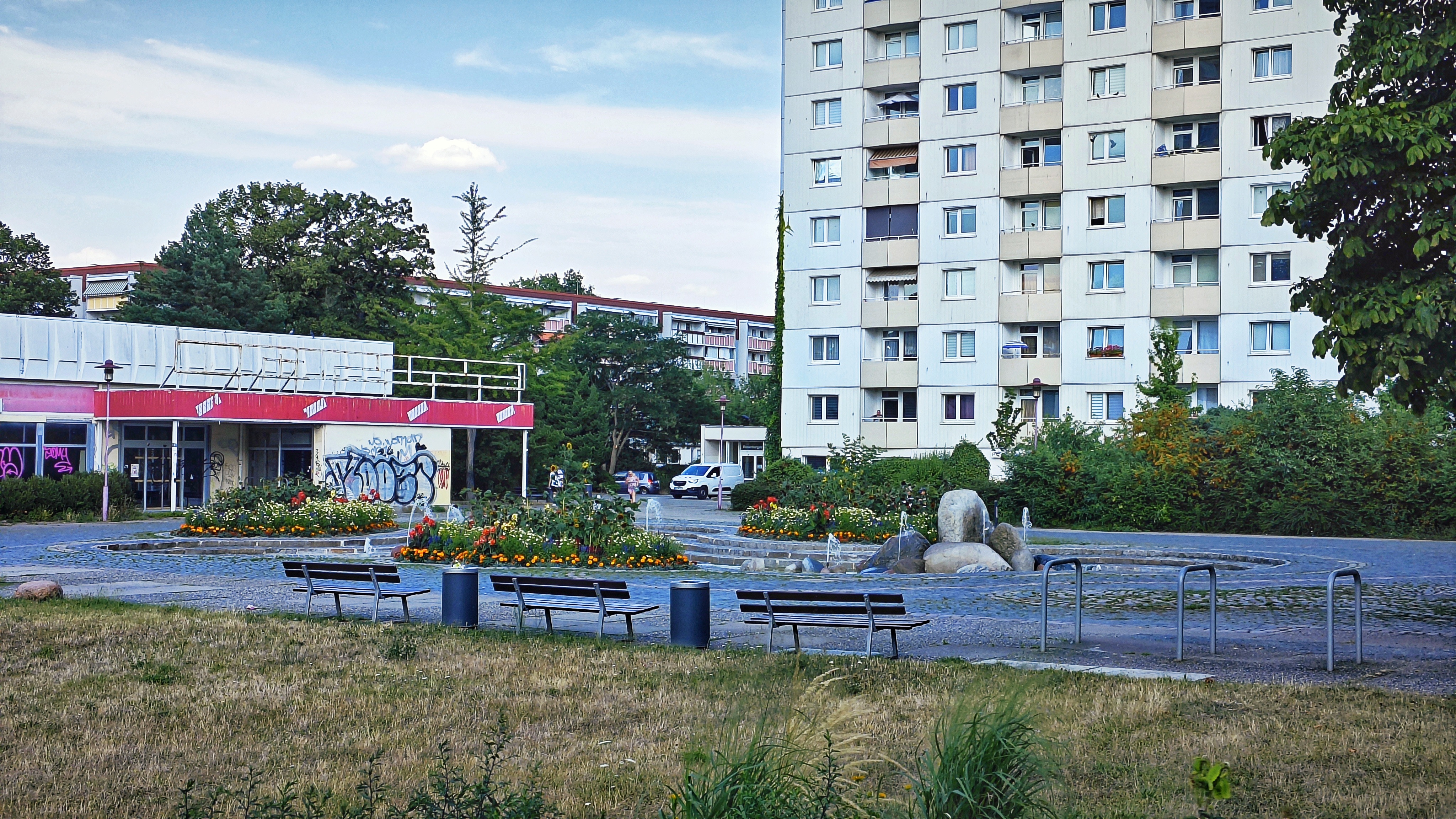 Ein schöner Brunnen, doch rundherum ansonsten nur Trostlosigkeit: der Flachbau eines Ladens, dessen Wände mit Graffiti tapeziert sind, ein vielstöckiges Hochhaus und von der Sonne versengtes Gras.